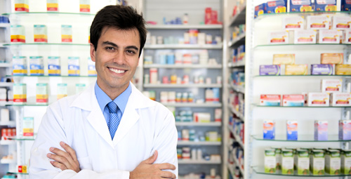 A pharmacist stood behind his counter with his arms folded staring out to the camera